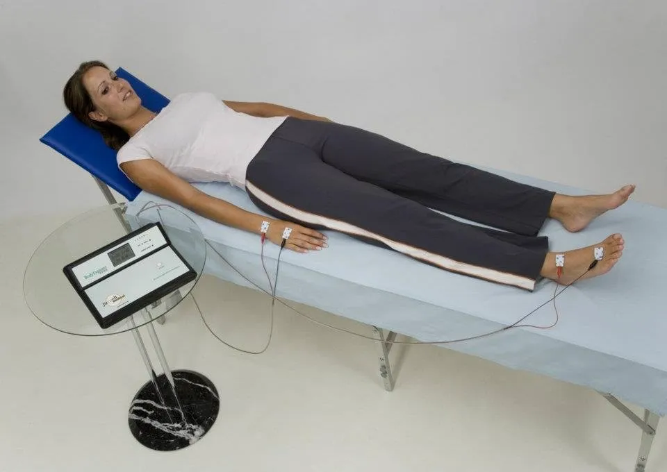 Pomiar składu ciała metodą bioimpedancji — pacjentka leży z elektrodami na dłoni i stopie, obok analizator Body Explorer